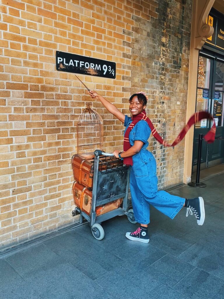Abigail at Platform 9 3/4 with one hand on a trolley and another hand holding a wand up at the plaque 